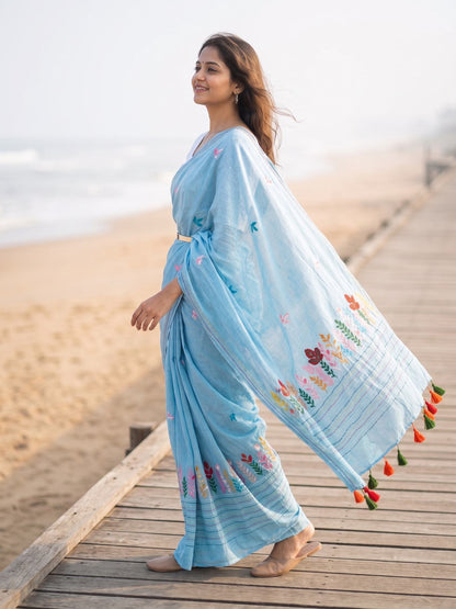 Woman draped in sky blue Belpotra mul cotton saree with hand-embroidered bael leaf motifs, walking on a beach boardwalk